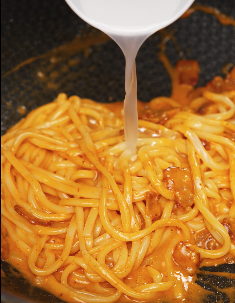 Pasta water being poured into a pan of gochujang carbonara noodles to loosen the spicy sauce. The thick udon noodles are coated in a vibrant orange gochujang-based mixture, blending Korean heat with creamy carbonara textures.