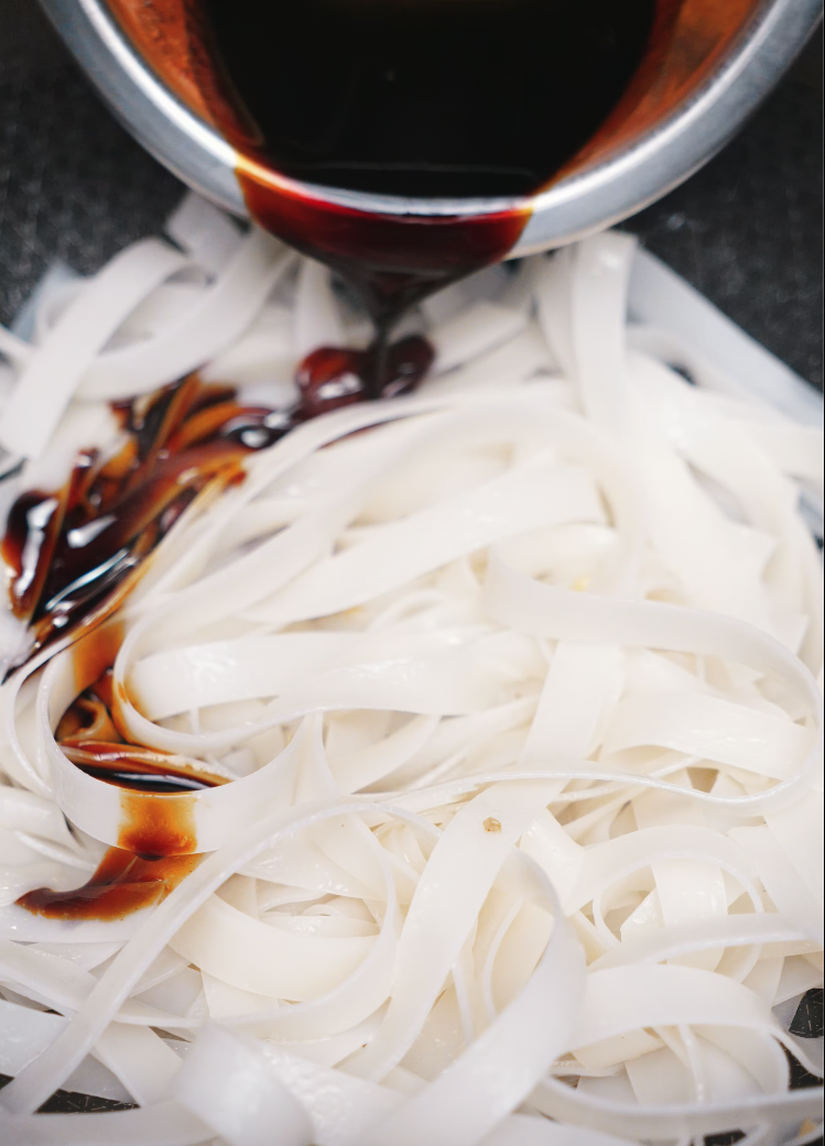 A savory blend of soy sauce being poured over freshly boiled rice noodles in preparation for Pad See Ew. This dark, umami-rich sauce forms the flavor foundation of the dish.
