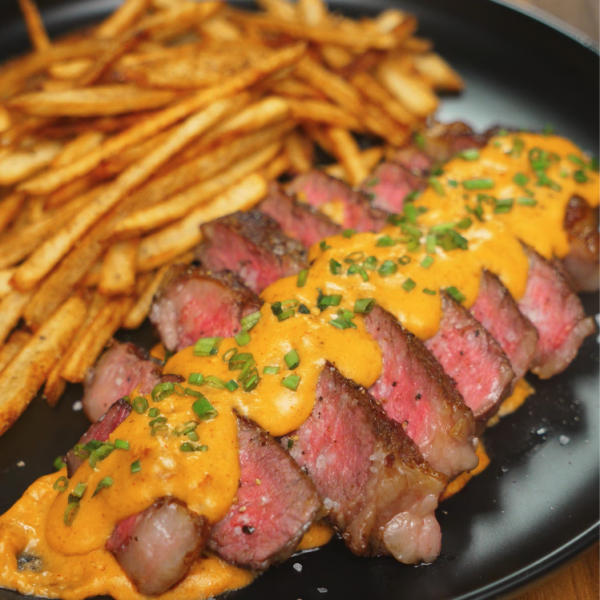 Plated ribeye steak slices topped with gochujang béchamel and chopped chives, alongside a generous portion of crispy five-spice fries.