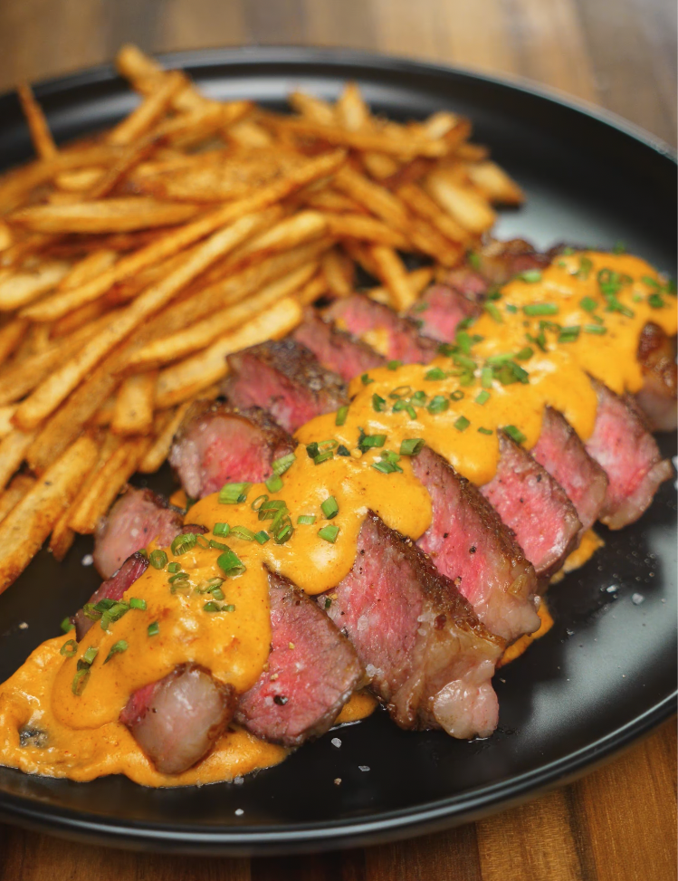 Plated ribeye steak slices topped with gochujang béchamel and chopped chives, alongside a generous portion of crispy five-spice fries.