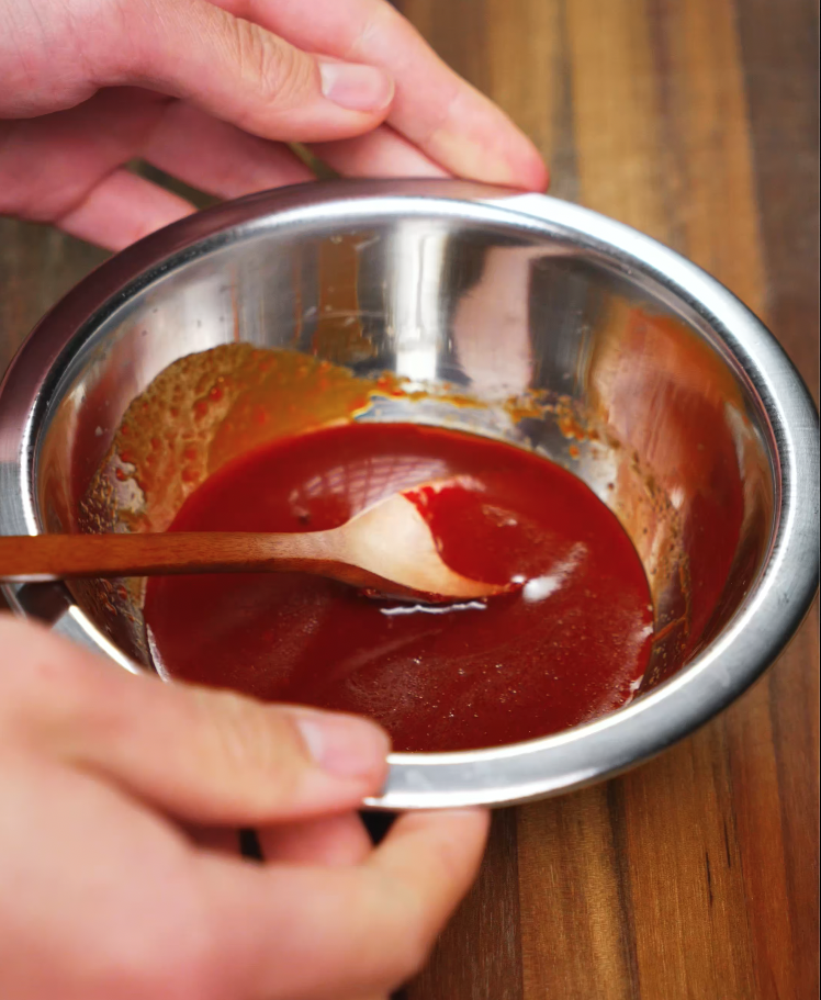 A hand holds a small metal bowl holding the gochujang glaze made out of gochuajng, soy sauce, sesame oil, rice wine vinegar, and honey for bbq (barbeque) gochujang chicken.