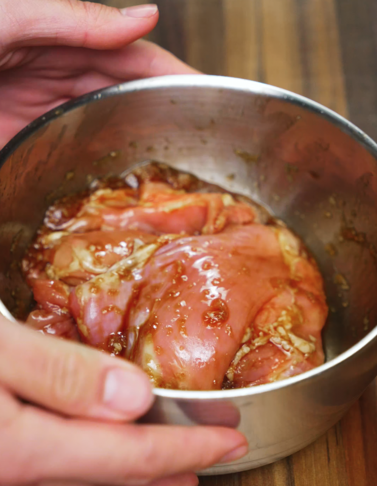 Hand holding mixing bowl filled with boneless skinless chicken thighs marinated in garlic, ginger, and soy sauce for 15 minutes for barbeque gochujang chicken