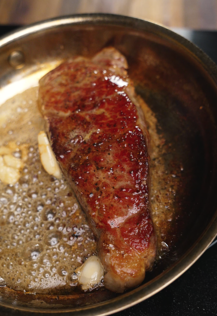 A thick-cut steak sizzles in a hot skillet with bubbling garlic-infused butter, developing a rich brown crust. The marbled meat is perfectly caramelized, capturing the start of a juicy and flavorful sear.