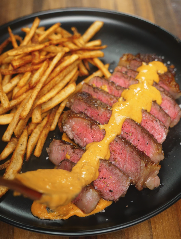 Medium-rare ribeye steak sliced and topped with creamy gochujang béchamel, served with a side of crispy five-spice seasoned fries on a black plate.