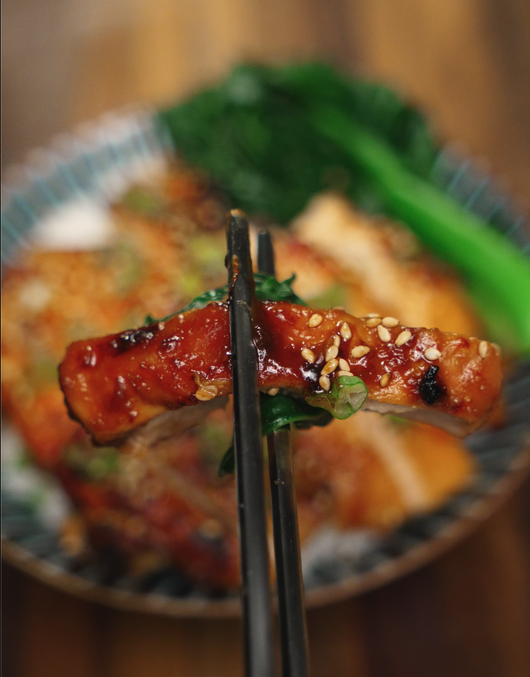 A glossy slice of miso-glazed chicken is held by chopsticks over a bed of rice and bok choy, revealing its tender and juicy interior coated with sesame seeds and scallions.