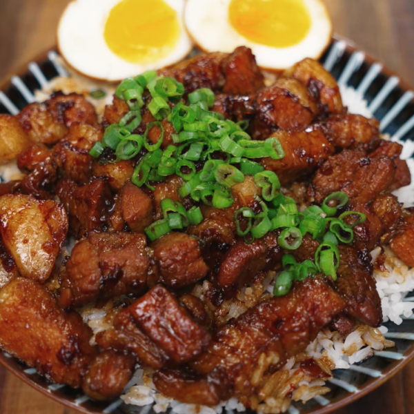 A full view of a soy braised pork belly rice bowl topped with generous slices of tender pork belly, vibrant green scallions, and halved soft-boiled eggs, all plated beautifully over a bed of white rice.