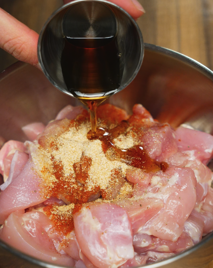 A shot of sesame oil or soy sauce being poured over raw, boneless chicken pieces seasoned with garlic powder, onion powder, paprika, and other spices, preparing for a flavor-packed marinade.