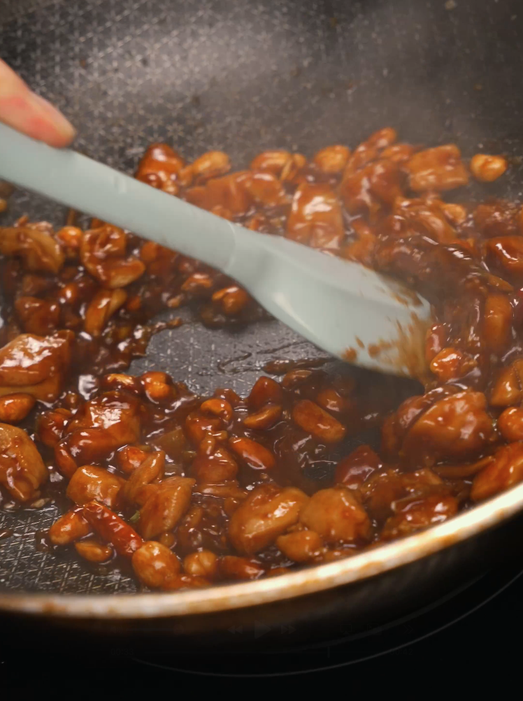 A nonstick pan filled with diced chicken and peanuts being coated in a glossy, dark Kung Pao sauce using a silicone spatula. The sauce is thick and sticky, clinging to every piece as it finishes reducing.