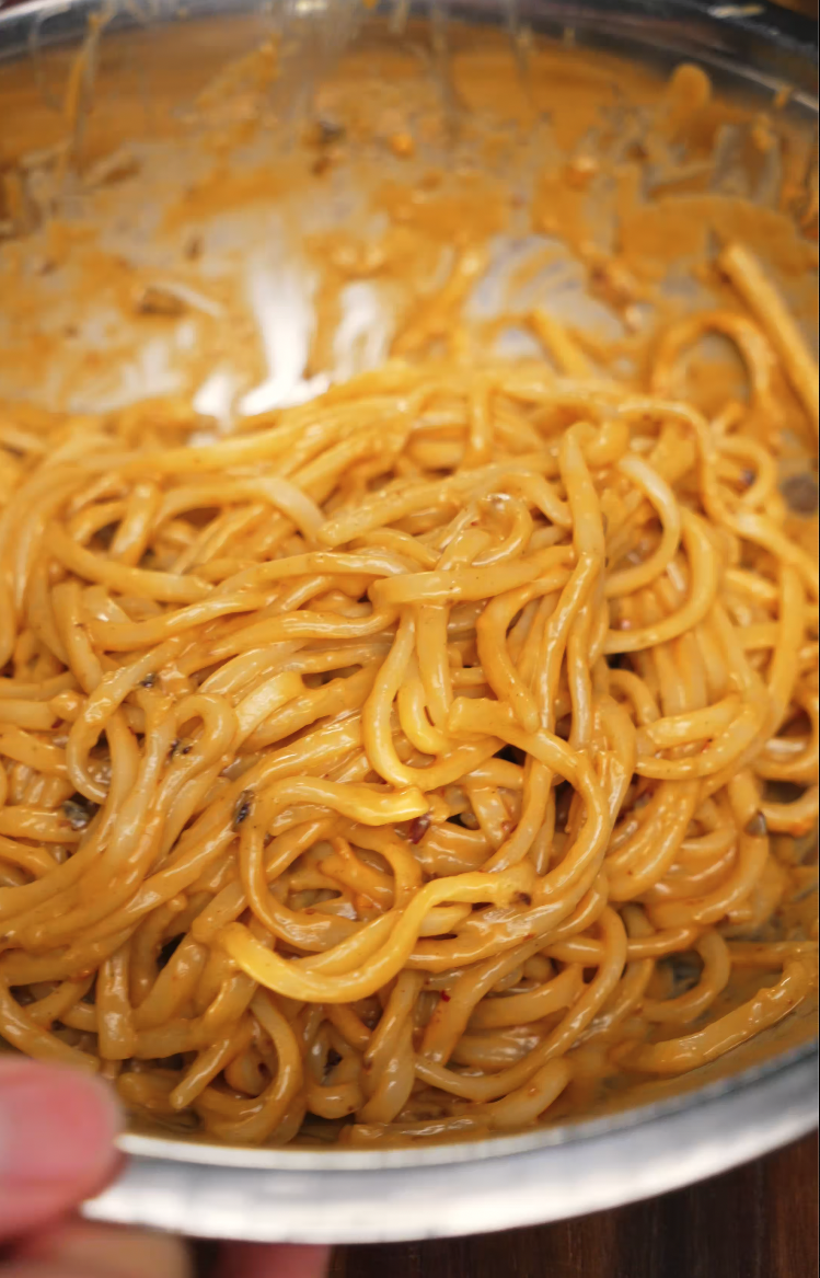 Close-up of freshly cooked noodles coated in a thick, creamy spicy peanut sauce inside a mixing bowl. The noodles are glossy and well-mixed, showing off the rich, nutty coating made from peanut butter and chili oil for maximum flavor.