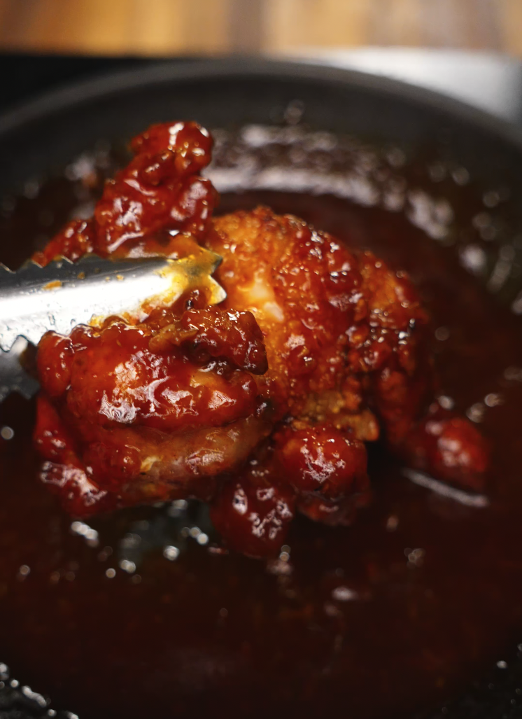 A glossy, spicy-sweet gochujang glaze coats a perfectly crispy piece of fried chicken held in tongs, just lifted from the bubbling sauce in the pan.