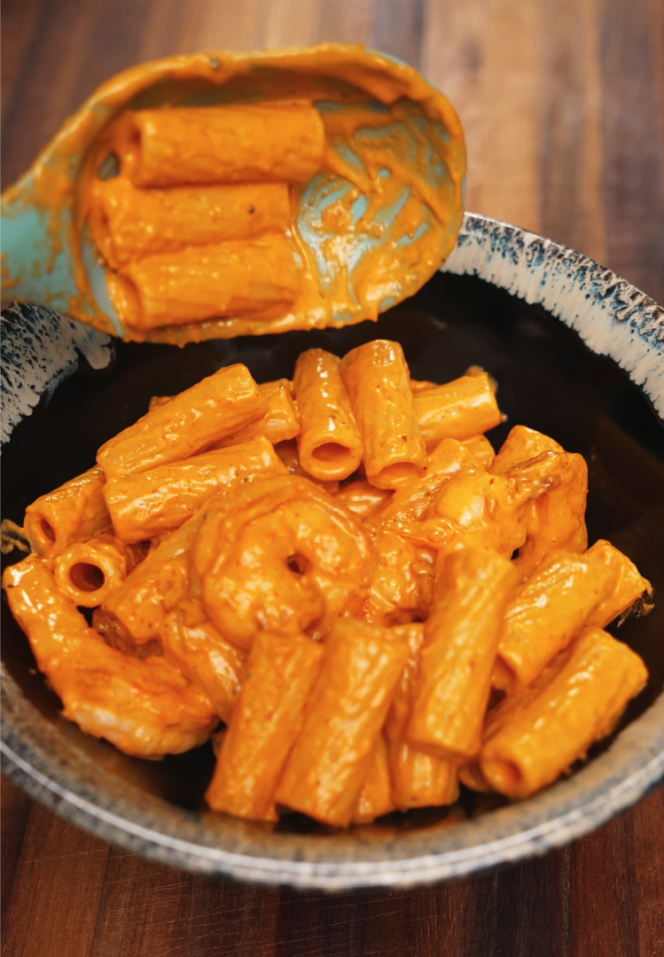 A spoonful of creamy rigatoni and shrimp is lifted over a bowl, coated in a rich, spicy orange sauce made with chili flakes, garlic, and cream. The glossy finish on the pasta highlights its velvety texture and bold flavor.