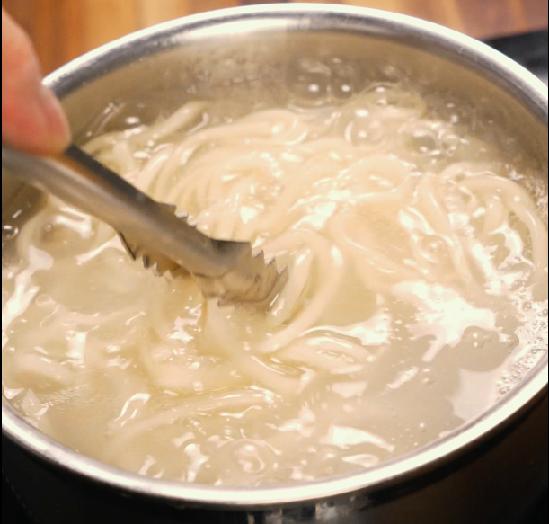 Noodles boiling in a pot of water being stirred with tongs — prepping the base for dan dan noodles.