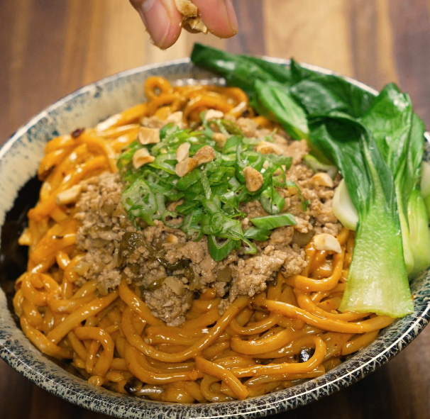 Hand sprinkling crushed peanuts onto a bowl of dan dan noodles topped with pork, scallions, and bok choy.