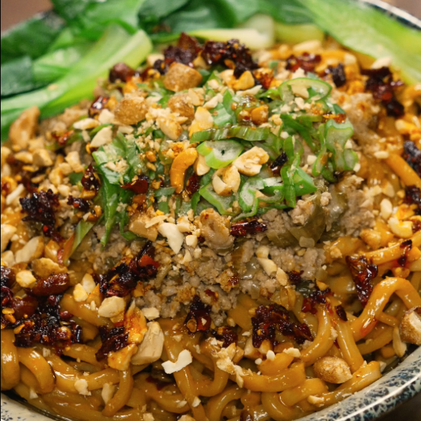 Full bowl of dan dan noodles garnished with scallions, peanuts, chili oil, and bok choy — spicy and savory Chinese noodle dish.