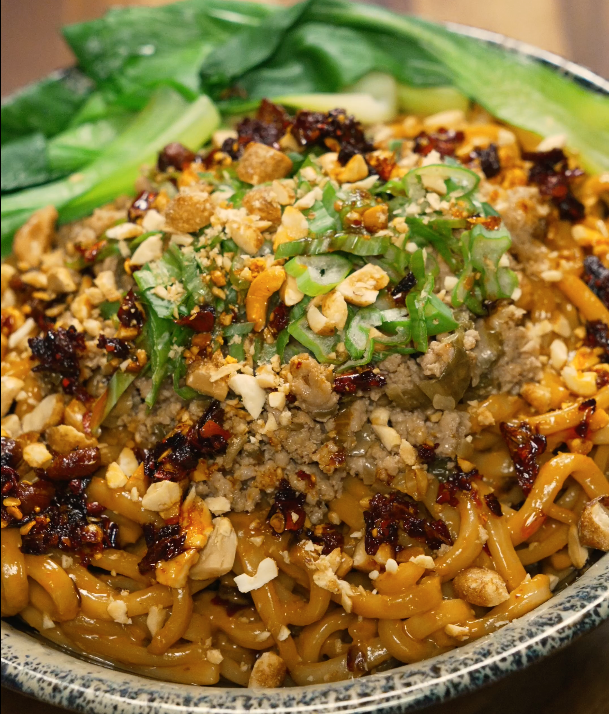 Full bowl of dan dan noodles garnished with scallions, peanuts, chili oil, and bok choy — spicy and savory Chinese noodle dish.