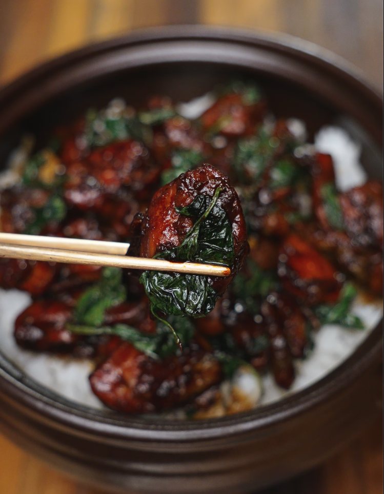 Close-up of a glossy, caramelized piece of 3 Cup Chicken lifted by chopsticks, with basil clinging to its surface. The glaze’s sheen and deep color highlight the richness and complexity of this Taiwanese favorite.