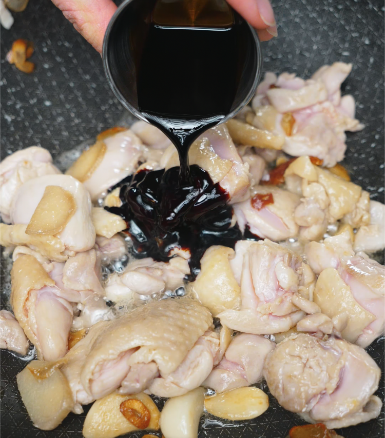 Pouring dark soy sauce into a skillet of pan-seared chicken and golden garlic cloves. This crucial step builds the bold, savory umami base for 3 Cup Chicken, as the soy sauce begins to sizzle and coat each piece of browned meat.