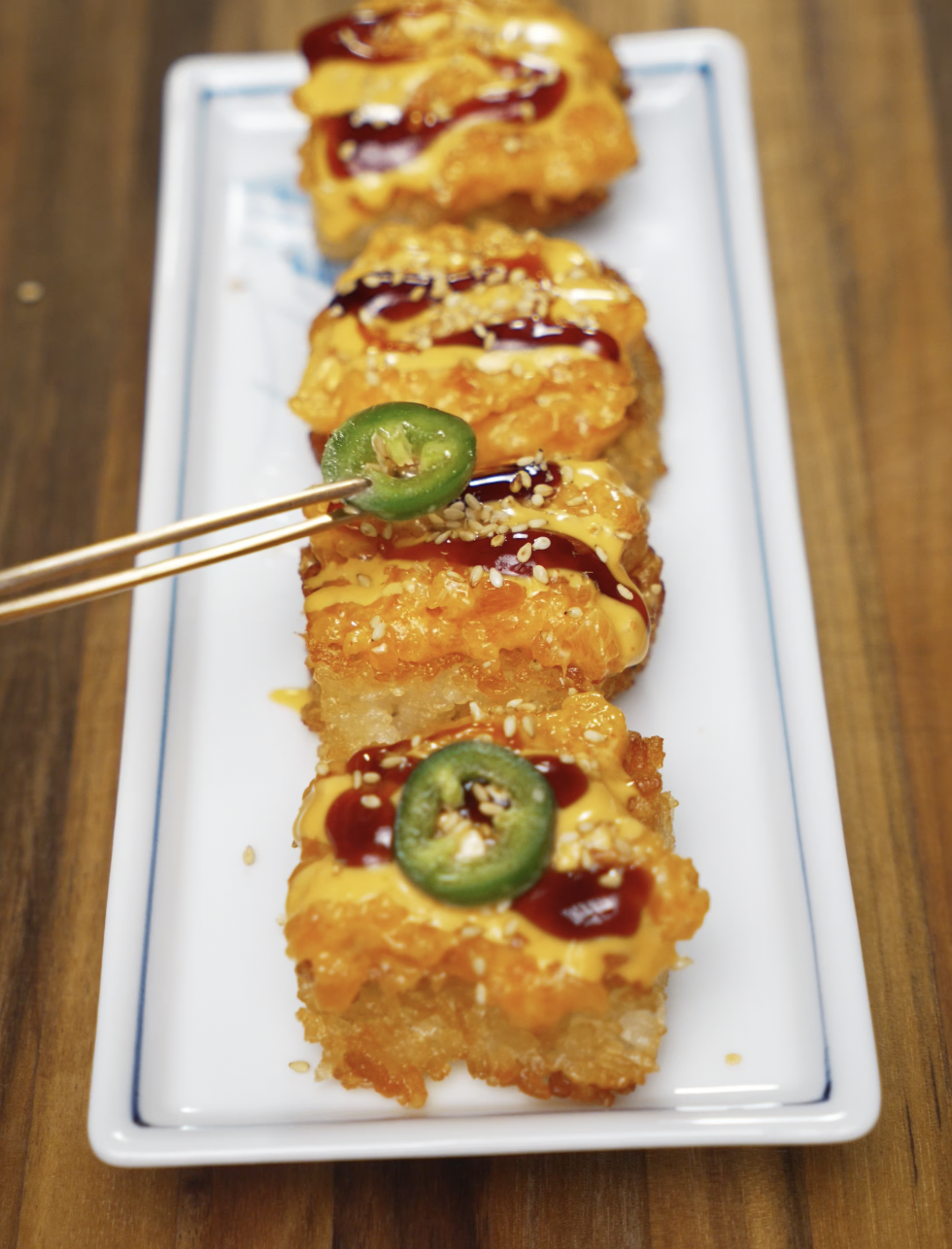 Chopsticks placing a slice of fresh jalapeño onto a golden, crispy salmon rice bite drizzled with spicy mayo and soy glaze, garnished with sesame seeds on a white platter.