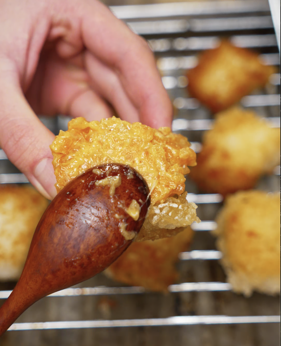 A hand holding a crispy golden rice cube while brushing spicy salmon on top with a wooden spoon. The rice is perfectly fried, showcasing its crunchy texture before assembling the salmon bites.