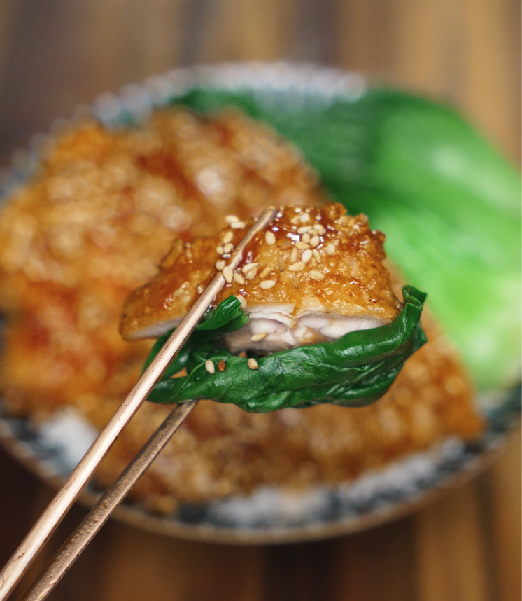 Chopsticks holding a bite of juicy soy garlic glazed chicken with toasted sesame seeds, wrapped in a tender leaf of bok choy for a perfect balance of savory and fresh flavors.