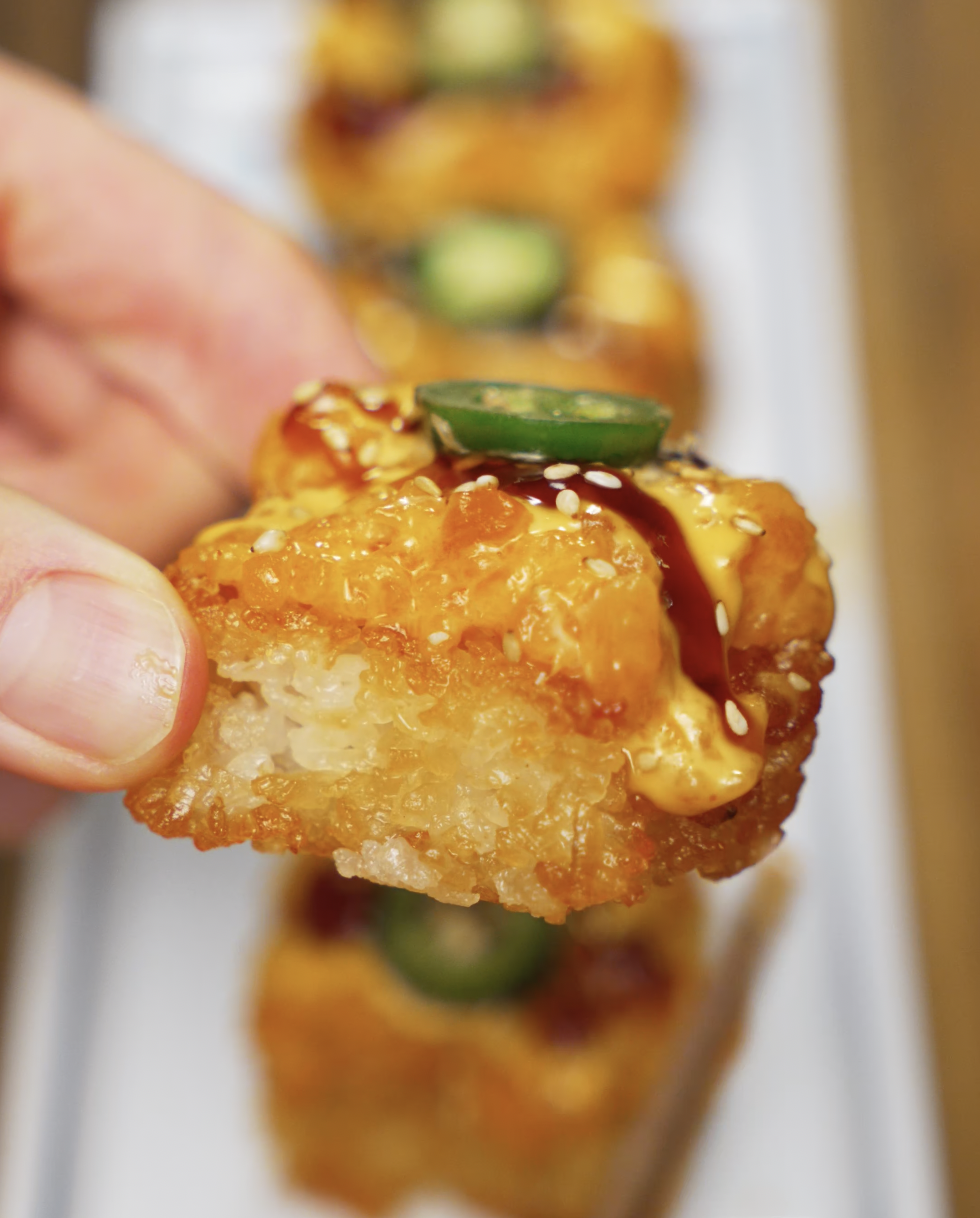 A hand holding a crispy rice bite topped with creamy, spicy salmon mixture, jalapeño, and sesame seeds. The texture of the crispy rice and the juicy salmon topping is clearly visible.