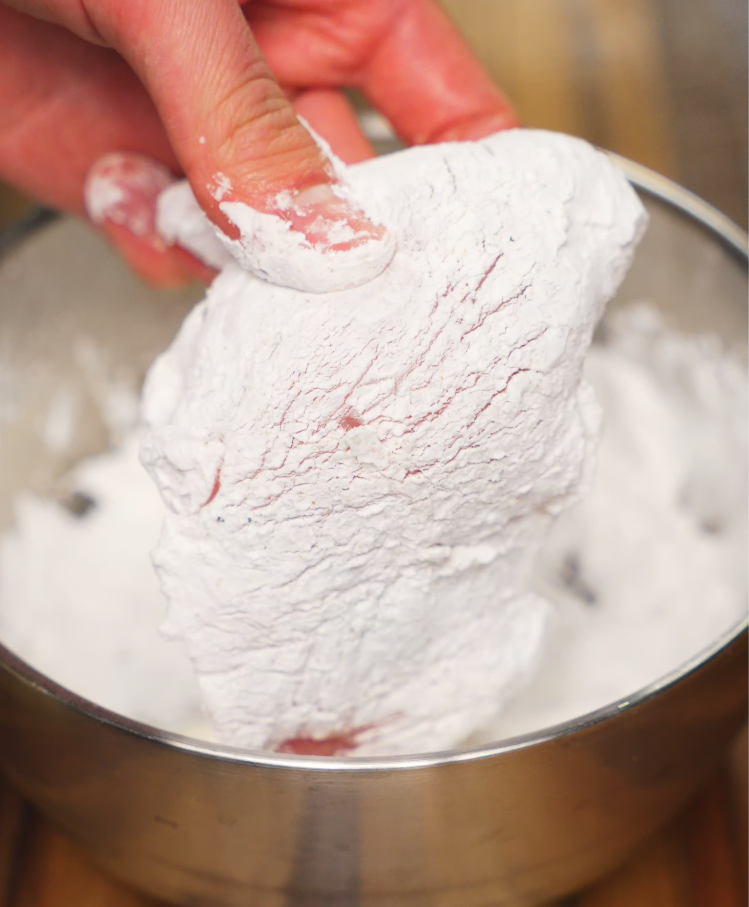 Close-up of raw chicken thigh being fully coated in a layer of cornstarch in a metal bowl. The starch will create a crispy outer layer when fried, essential for holding the soy garlic glaze.