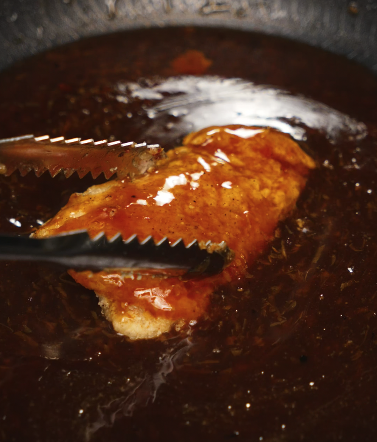 Crispy fried chicken thigh being glazed in a thick soy garlic sauce in a skillet. Tongs are used to flip and evenly coat the chicken, giving it a shiny, sticky texture.