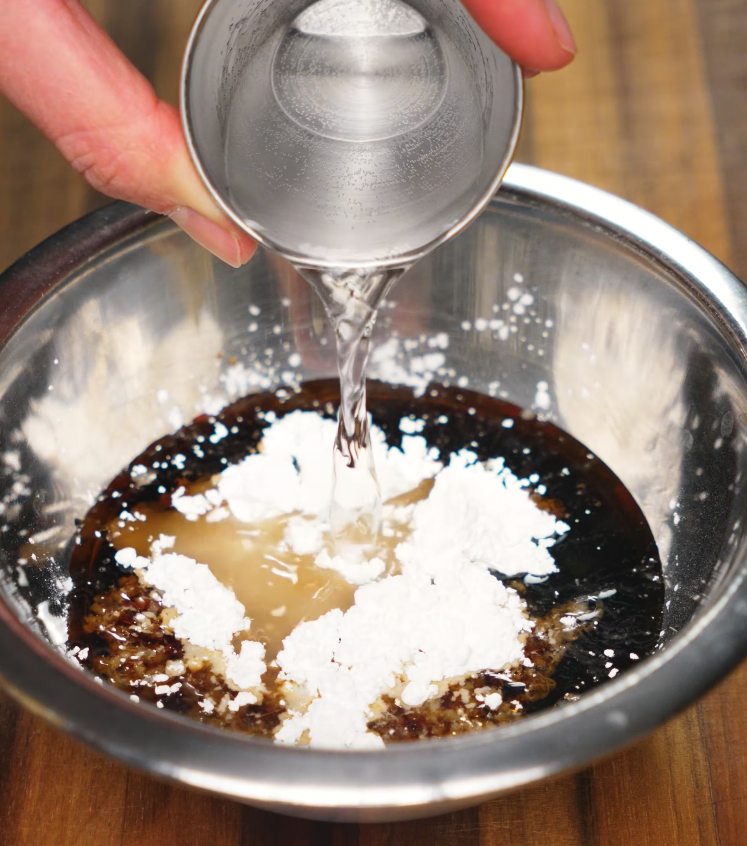 Cornstarch slurry being poured into a soy garlic sauce mixture in a metal bowl. The sauce includes soy sauce, sugar, and other Asian pantry staples and is thickened for glazing.