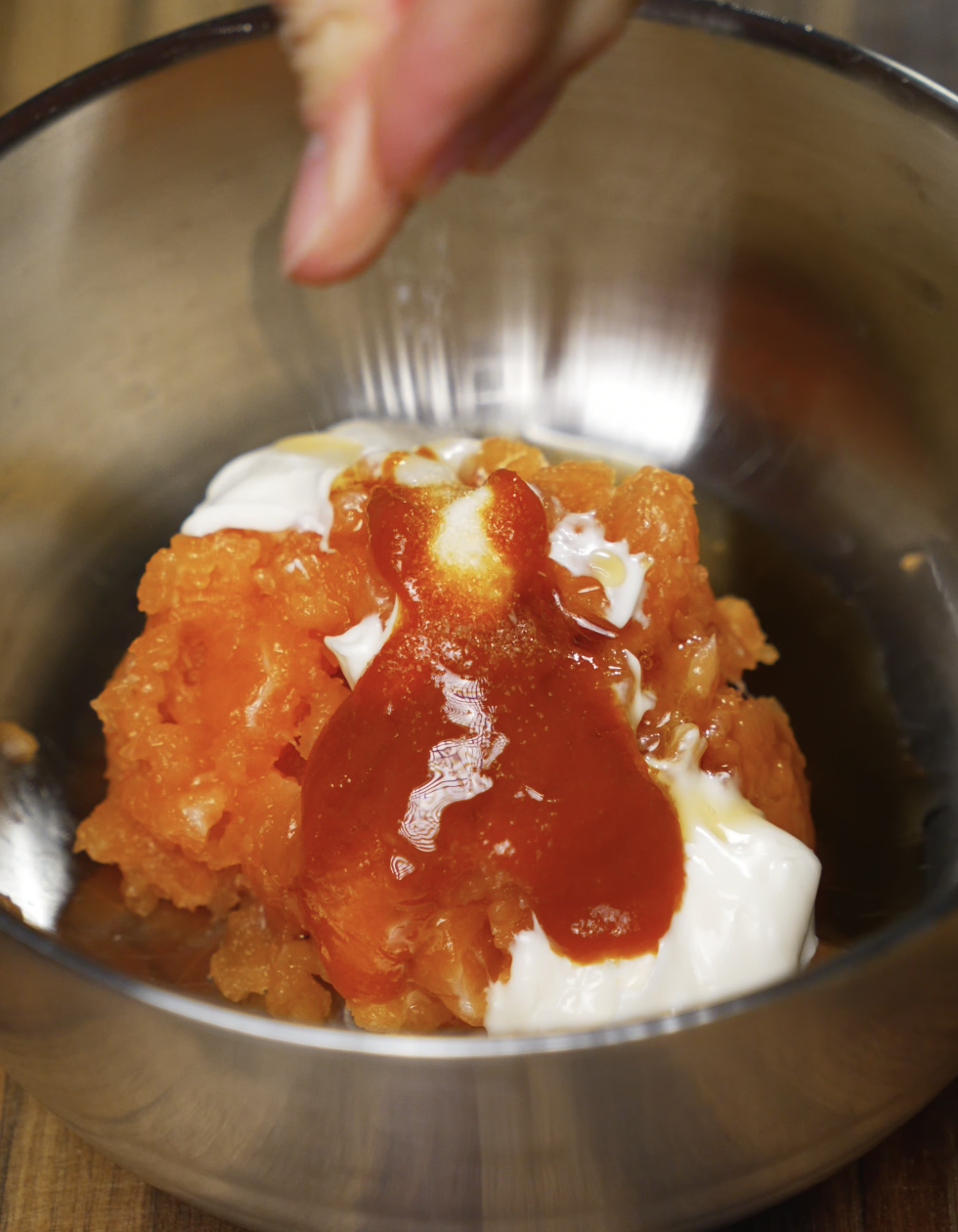 A metal bowl filled with chopped salmon, mayonnaise, and sriracha being seasoned with salt. This spicy creamy mixture will be used as a topping for crispy rice bites.
