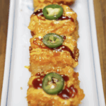 Rows of crispy rice squares topped with spicy salmon mix, spicy mayo, sweet soy glaze, and fresh jalapeño slices. Finished with sesame seeds and plated on a rectangular white dish.