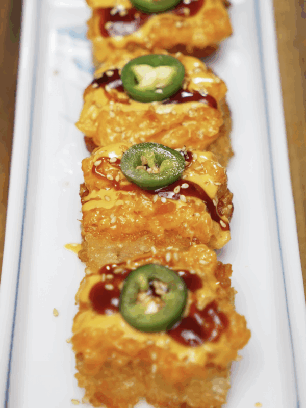 Rows of crispy rice squares topped with spicy salmon mix, spicy mayo, sweet soy glaze, and fresh jalapeño slices. Finished with sesame seeds and plated on a rectangular white dish.