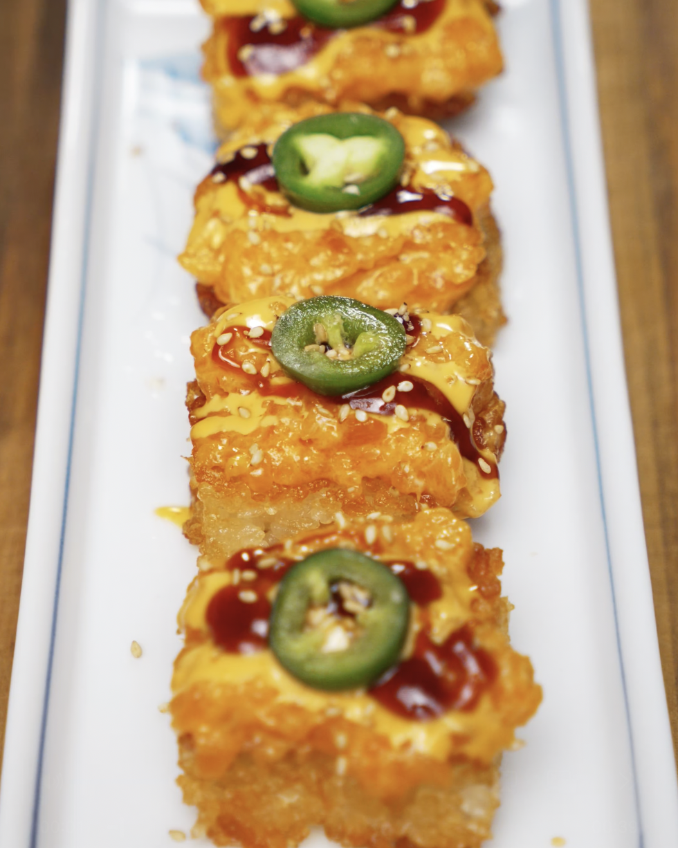 Rows of crispy rice squares topped with spicy salmon mix, spicy mayo, sweet soy glaze, and fresh jalapeño slices. Finished with sesame seeds and plated on a rectangular white dish.