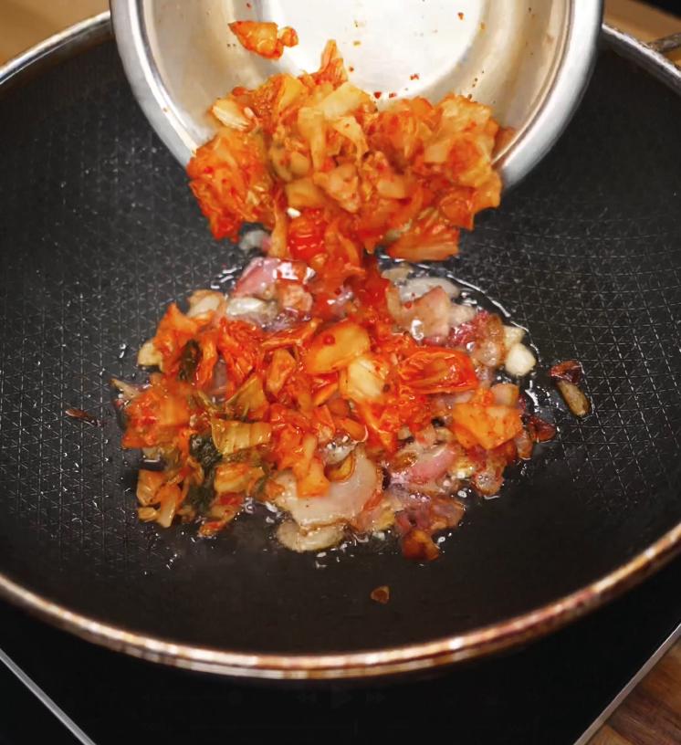 Chopped kimchi and rendered bacon being added to a pan with hot oil to begin the stir-fry base.