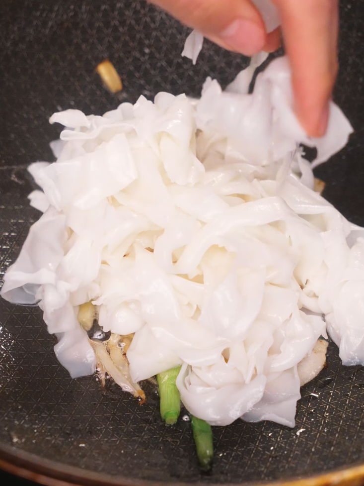 Fresh wide rice noodles being added into a wok with stir-fried garlic and green onions to start the stir-fry for Beef Chow Fun. This step sets the foundation for the noodle’s texture and flavor.