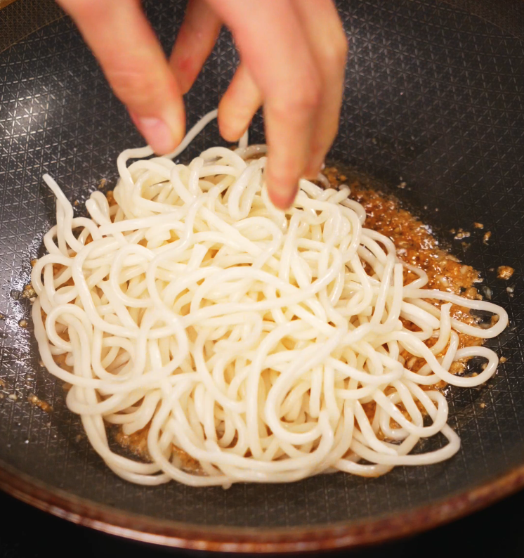 Fresh udon noodles being added to a garlic butter sauce in a frying pan, ready to soak up the savory flavors.