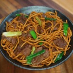 A finished bowl of beef lo mein featuring perfectly coated noodles, tender slices of beef, and vibrant green onions. This final presentation showcases the glossy sauce and balanced stir-fry technique.