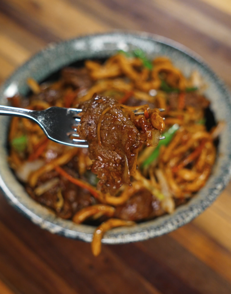A close-up of tender slices of seared beef lifted from a bowl of beef yaki udon, highlighting the glossy sauce-coated noodles, onions, and vibrant vegetables.
