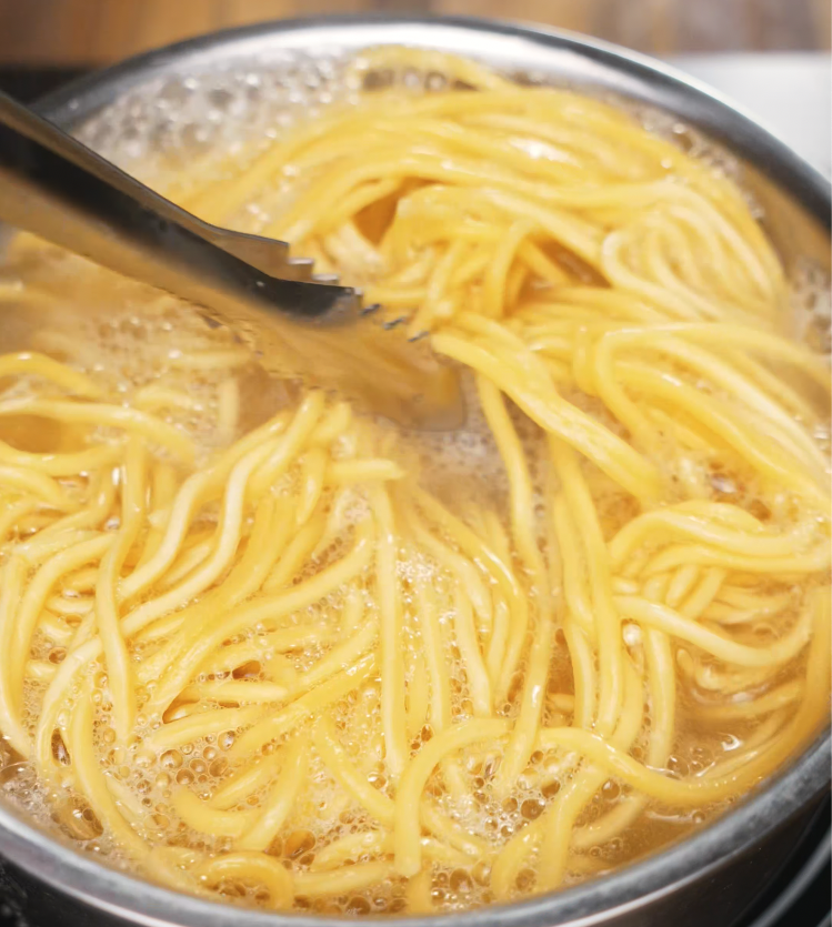 Lo mein noodles boiling in a pot of water with metal tongs lifting the strands. Cooking until tender and chewy for the perfect noodle texture.