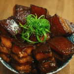 A close-up of glossy braised pork belly stacked in a bowl and topped with thinly sliced scallions, highlighting the caramelized glaze and tender texture.
