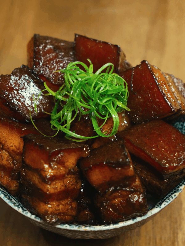 A close-up of glossy braised pork belly stacked in a bowl and topped with thinly sliced scallions, highlighting the caramelized glaze and tender texture.