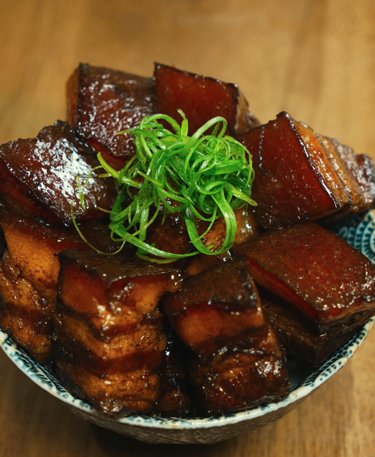 A close-up of glossy braised pork belly stacked in a bowl and topped with thinly sliced scallions, highlighting the caramelized glaze and tender texture.