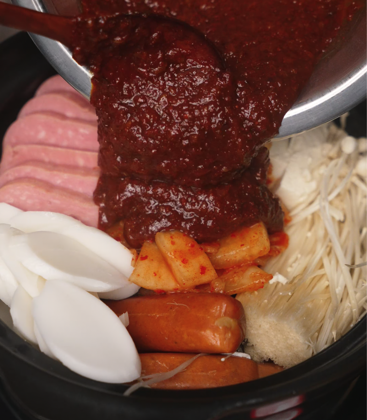 Pouring rich, red gochujang soup base over neatly arranged kimchi, sausages, and rice cakes before simmering.