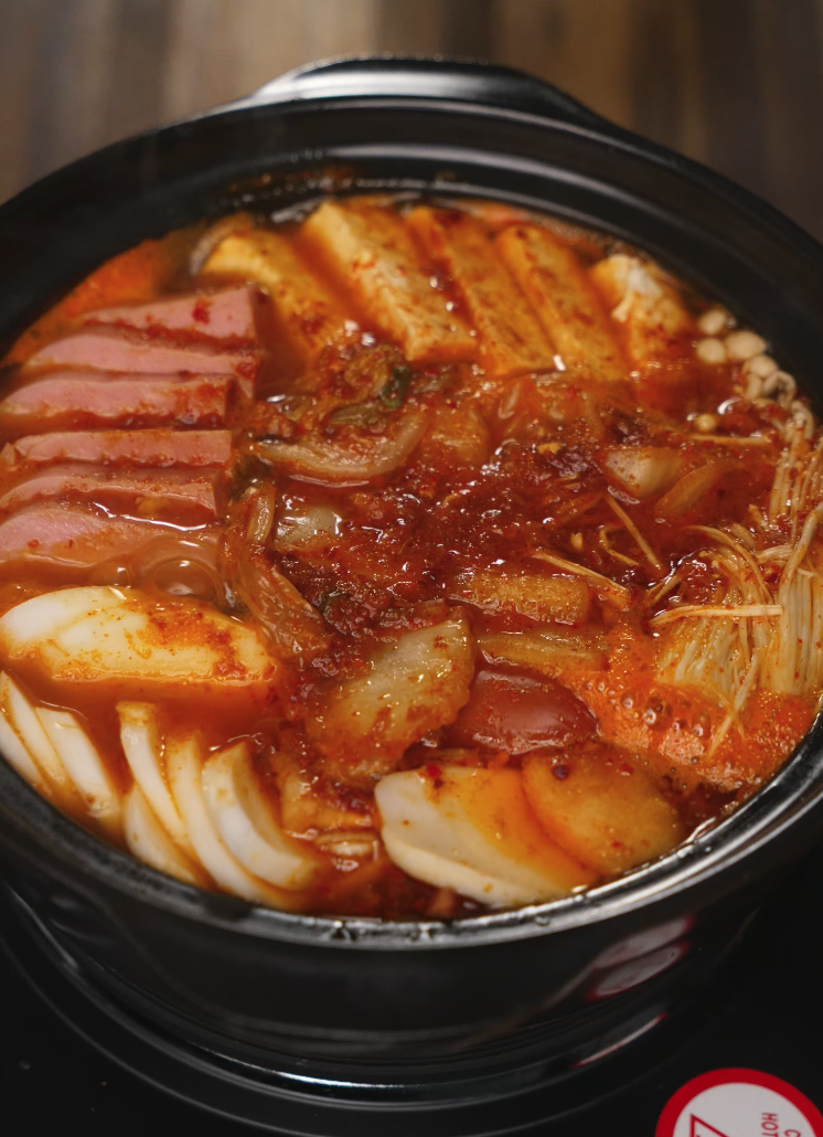 Korean army stew bubbling in a pot with visible spam, rice cakes, kimchi, mushrooms, and tofu soaking in spicy broth.
