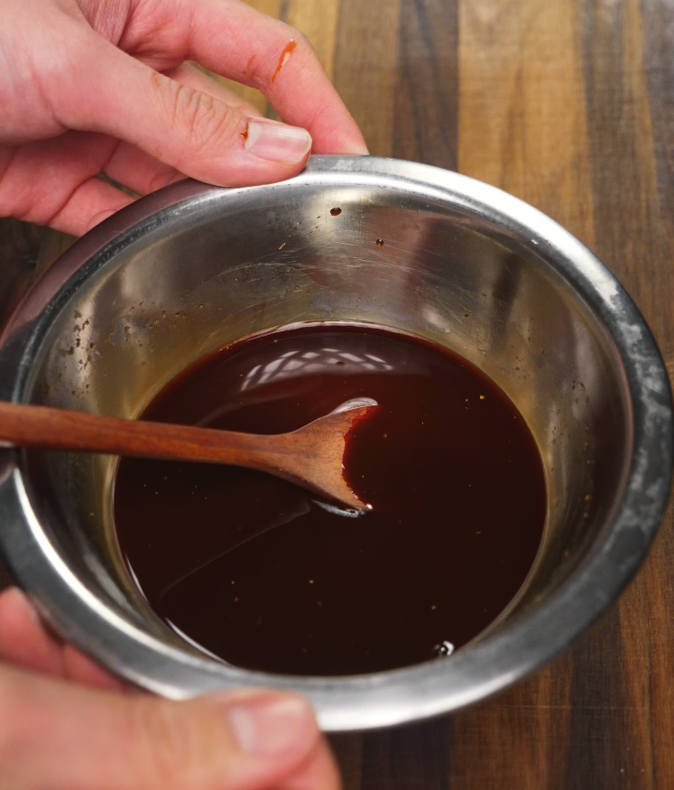 Overhead shot of rich brown lo mein stir-fry sauce in a metal bowl, stirred with a wooden spoon, ready to coat the noodles with savory, umami-packed flavor.