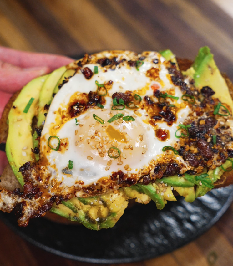 Close-up of one slice of chili oil egg toast held in hand, showing perfectly cooked yolk, creamy avocado, and crispy chili topping.