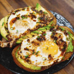 Close-up of two slices of sourdough toast topped with avocado, sunny side up eggs, and chili crisp, served on a black plate.