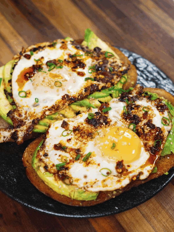 Close-up of two slices of sourdough toast topped with avocado, sunny side up eggs, and chili crisp, served on a black plate.