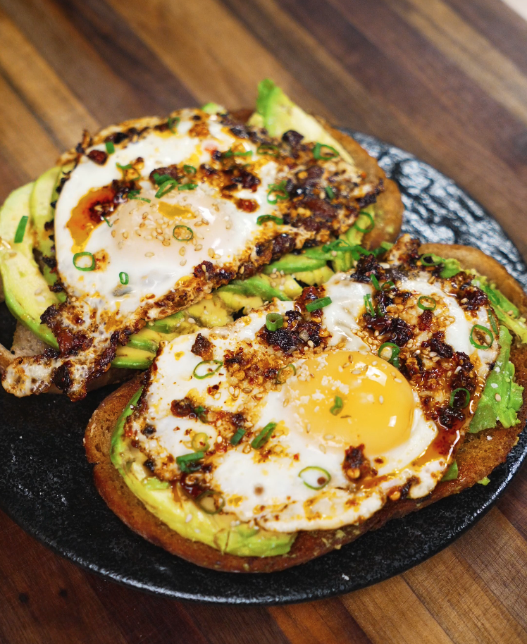 Close-up of two slices of sourdough toast topped with avocado, sunny side up eggs, and chili crisp, served on a black plate.