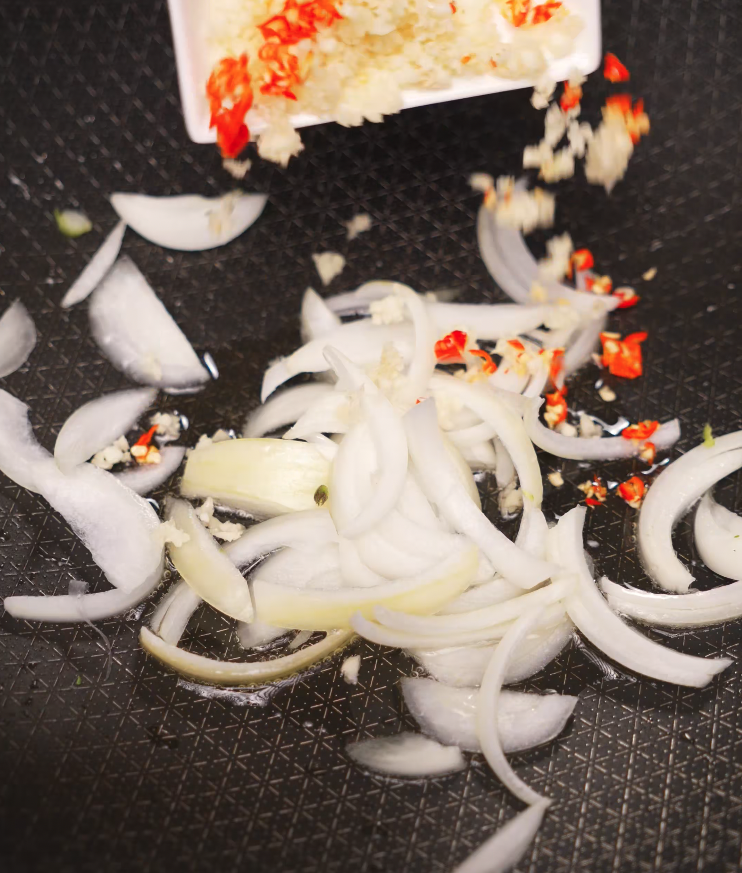 Sliced onions, minced garlic, and chopped red chilies being tossed into a sizzling pan of hot oil, creating the fragrant base for the stir-fry.