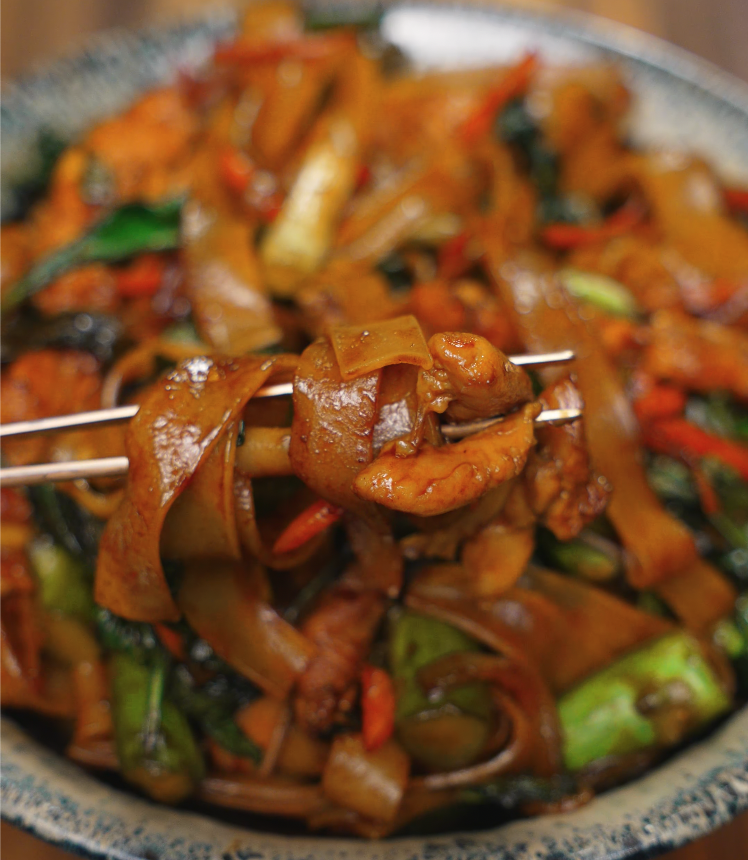 Chopsticks lifting a perfect bite of drunken noodles with glossy noodles, tender chicken, and vegetables from the serving bowl.